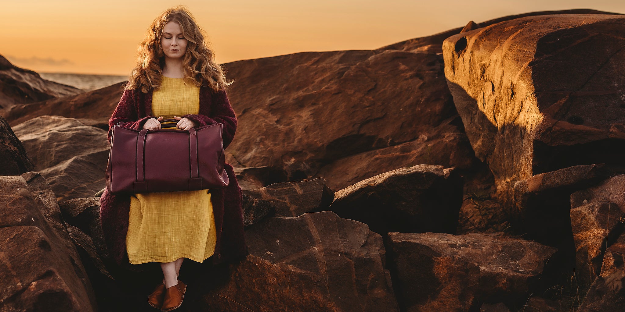 Tyttö istuu kalliolla violetti matkalaukku sylissään.